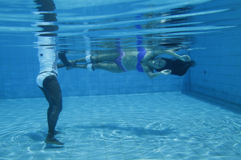 A person swimming in the water with another person standing on one side.